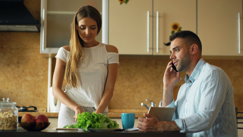 Couple in a kitchen, one partner distracted by a phone during conversation
